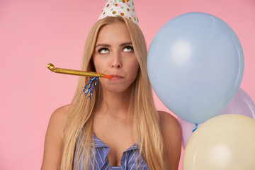 Bored pretty blonde woman with long hair rolling eyes and keeping party horn in her mouth,...