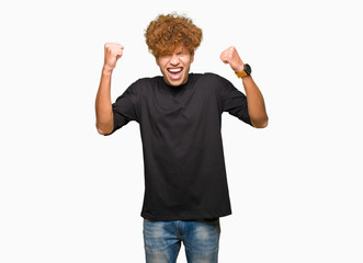 Young handsome man with afro hair wearing black t-shirt very happy and excited doing winner gesture with arms raised, smiling and screaming for success. Celebration concept.