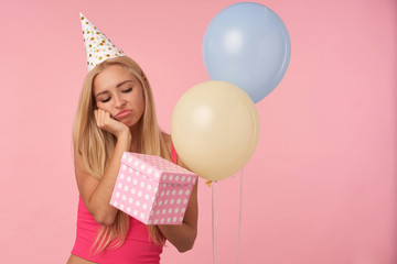 Indoor shot of sad young blonde woman in holiday outfit keeping gift-wrapped box, keeping chin on raised hand and looking at box with disappointment, standing over pink background and helium balloons