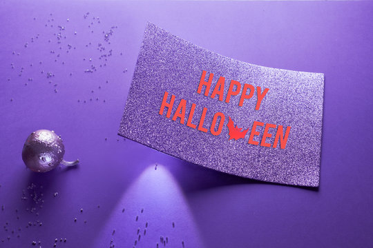 Spotlight shining from underneath glittering paper card with text "Happy Halloween". Creative purple Halloween background with glittering pumpkin painted metallic on neon paper.