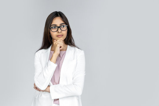 Thoughtful doubtful young adult indian businesswoman in formal wear white suit stand isolated on grey background with copy space. Serious girl in eyewear looking away doubting feel confused uncertain