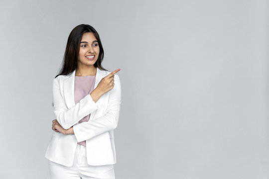 Smiling young adult indian business lady in formal white suit looking away pointing index finger aside. Happy woman professional saleswoman customer standing isolated on grey background with copyspace