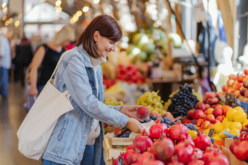 Woman is chooses fruits and vegetables at food market. Reusable eco bag for shopping. Sustainable lifestyle. Eco friendly concept.