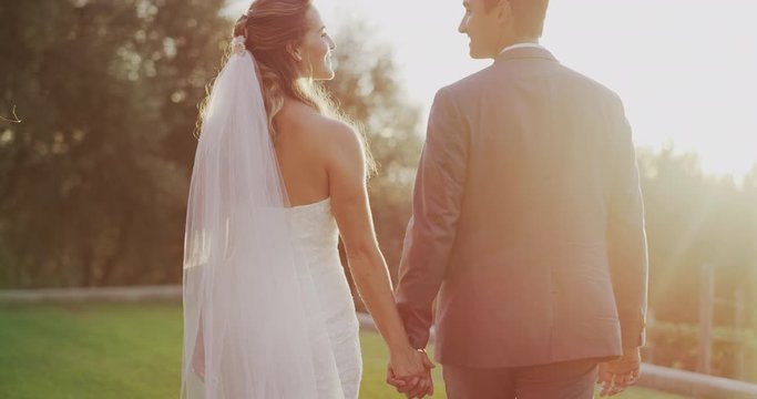 Happy diverse bride and groom holding hands and smiling at each other walking together at sunset on their wedding day
