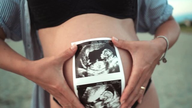 Close-up shot of beautiful tiny woman's belly and sonogram photo, pregnant woman dancing in sunset and posing on camera. Ultrasound examination, photo of unborn baby child. Happy future mother dancing
