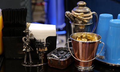 At the bar: black tubes, napkins, blueberries in a copper plate, blue cups, a metal figurine, a bucket of physalis, toothpicks, a silver teapot