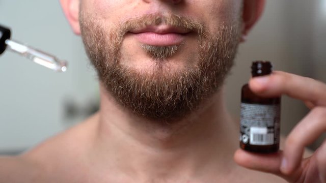 Handsome bearded man staying in the bathroom in front of mirror and holding opened glass bottle with fresh aromatic essential oil for softening dry man's beard after shaving and irritation. Barber's