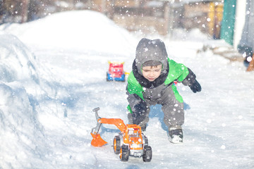 Fototapeta na wymiar Portrait of cute little toddler sitting on snow and playing with his yellow tractor toy in the park. Child playing outdoors. Happy boy with construction toy. Lifestyle concept
