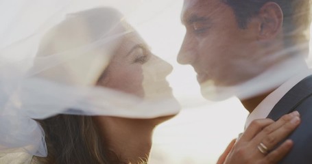 Intimate close up shot of a multi ethnic bride and groom smiling and looking at one and other underneath a white veil, intimate marriage ceremony at sunset