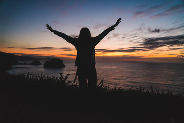 Back of a young traveler girl standing on on the edge of a cliff by the ocean coast watching a beautiful sunset. Arms wide open. Freedom concept