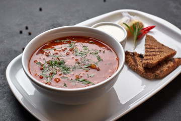 Red borscht on a white plate with bread and sour cream, on a dark background