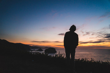 Back of a young traveler standing on on the edge of a hill by the ocean coast watching a beautiful sunset.