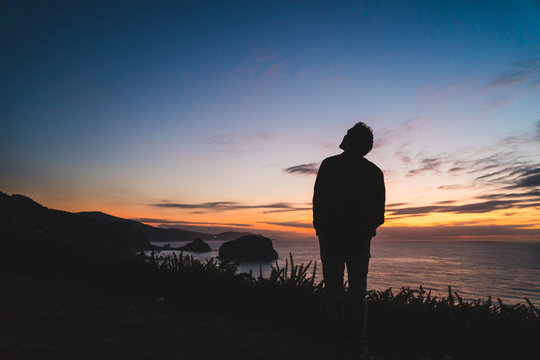 Back of a young traveler standing on on the edge of a hill by the ocean coast watching a beautiful sunset.