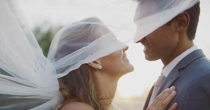 Intimate close up shot of multi ethnic bride and groom looking into each others eyes and smiling beneath a white veil, multi ethnic wedding ceremony moment at sunset with golden sun flare