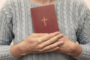 Young praying christian woman's hands holding holy bible with a cross on a cover