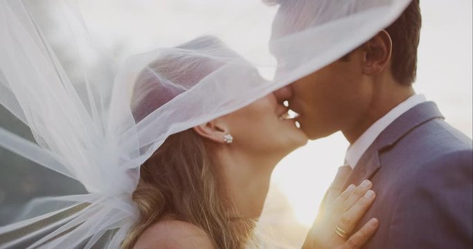 Intimate close up shot of multi ethnic bride and groom looking into each others eyes and kissing beneath a white veil, multi ethnic wedding ceremony moment at sunset with golden sun flare