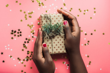Black girl is holding a gift of Kraft paper on a pink background with confetti. Womans hands...