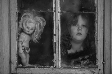 Portrait of lonely little child with an old doll looking out of window with relections