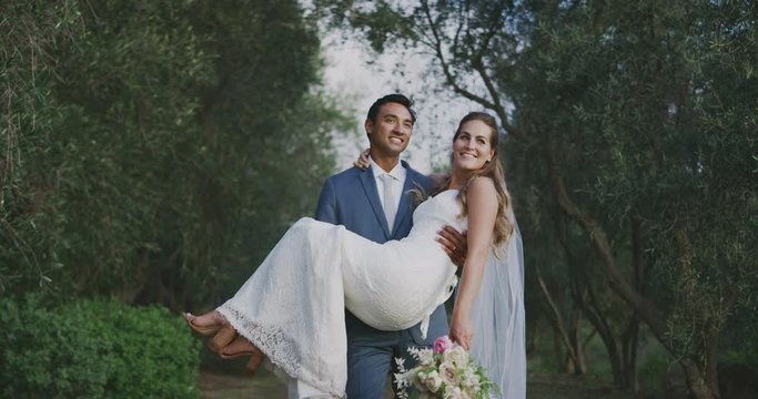 Happy multi ethnic groom carrying his bride smiling at her in nature on their wedding day, happy diverse couple getting married