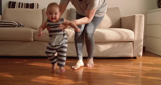 baby first step Slow motion of 6 months baby learning to walk at home mother holding baby arm walking on the floor indoor with sunlight coming family with baby lifestyles
