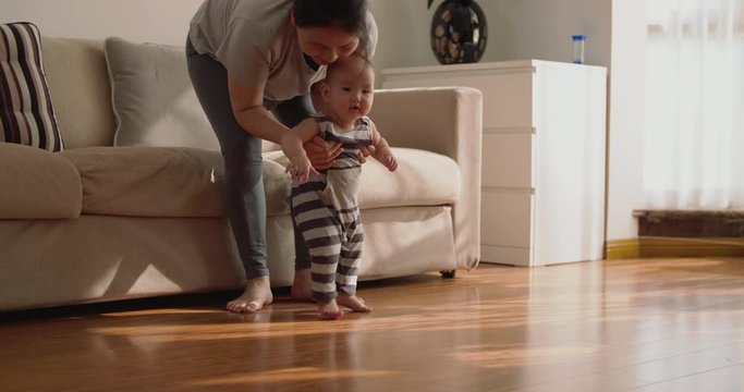 baby first step Slow motion of 6 months baby learning to walk at home mother holding baby arm walking on the floor indoor with sunlight coming family with baby lifestyles