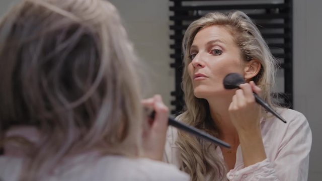 Young woman using a contour brush to apply make up to her face in a clean white bathroom in her own home or apartment preparing for the day with her morning routine
