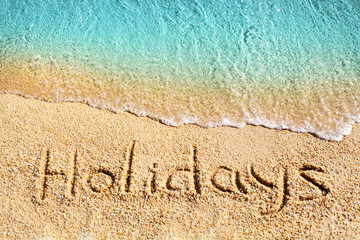 Word HOLIDAYS written on yellow sand, blue sea wave, white foam top view close up, handwriting letters text, ocean water, summer vacations on paradise island concept, tropical beach banner, copy space