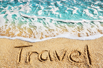 Word TRAVEL written on yellow sand, blue sea wave, white foam top view close up, handwriting...
