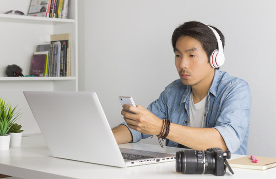 Asian Freelance Videographer Checking Multimedia File By Smartphone With Laptop In Home Office