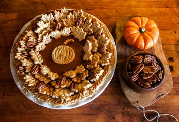 Homemade Pumpkin Pie Decorated with Pastry Cutouts of Autumn leaves with a center pumpkin, set on a wooden board with fresh pumpkins and pecans, selective focus