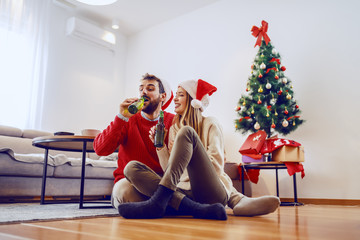 Adorable good-looking Caucasian couple with santa hats on heads sitting on floor in living room and drinking beer. In background is christmas tree with presents.