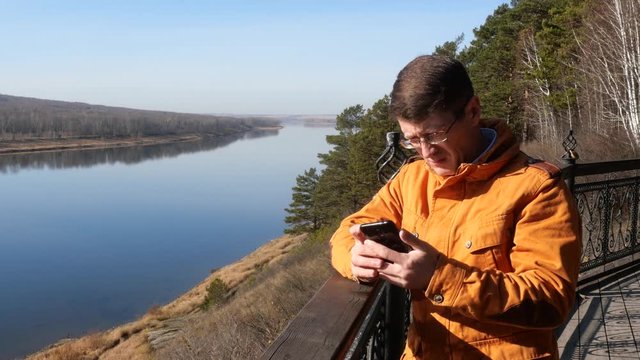 A man stands on the background of the river leaning on the railing and looking at the phone.