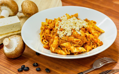 Fresh cooked vegetarian pasta noodles with red sauce, tofu on white plate and fresh mushrooms aside, on wooden table with fork and black soybeans. Side view.