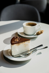 A souffle cake with chocolate on a white plate, fork, knife. A white cup of black coffee on a saucer, a cookie. Bright direct sun light. Vertical