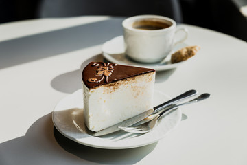 A souffle cake with chocolate on a white plate, fork, knife. A white cup of black coffee on a saucer, a cookie. Bright direct sun light. Horizontal
