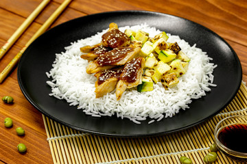 Fresh rice with heura - vegetable protein, teriyaki sauce, grilled zucchini on black plate, with chopsticks, green soybeans and soy sauce around, on wooden table. Healthy food.