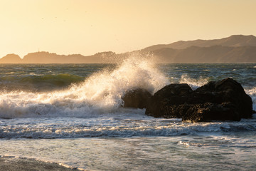 Fototapeta na wymiar Big wave from hard wind touching big rock of Baker Beach at sunset