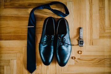 Stylish men's lacquered black shoes, tie, watch, wedding two rings lying on a light brown table. Wedding accessories groom on rustic wooden background. flat lay. top view.