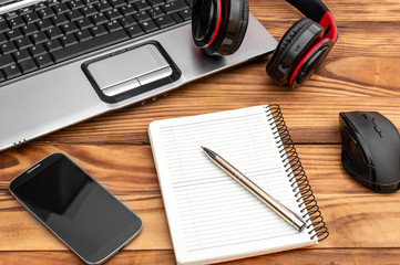 Wooden desk with blank notepad, laptop, smartphone, computer mouse and headphones.
