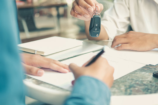 Dealer Salesman Giving Car Key To Owner. Client Signing Insurance Document Or Rental Car Lease Form