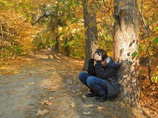 Upset problem child with head in hands sitting under tree in autumn park. Bullying, depression stress or frustration concept. 