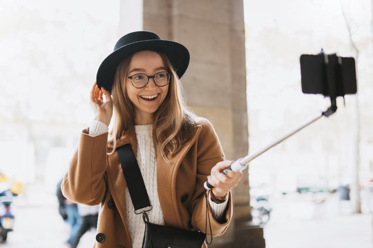 Smiling tourist girl taking photo selfie on smartphone mobile. Blogger hipster travels in Barcelona. Holiday concept in street. Traveler in glasses and hat self cellphone technology in europe trip