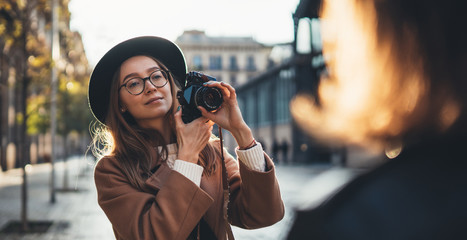 Fototapeta na wymiar Photographer in glasses with retro camera take photoshoot model girlfriend. Tourist smile girl in hat travels in Barcelona holiday with traveler friend. Sunlight flare street in europe