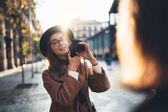 Photographer in glasses with retro camera take photo girlfriend. Tourist smiling girl in hat travels in Barcelona holiday with friend. Sunlight street in europe city. Traveler hipster shoot girls