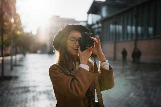 Outdoor smiling lifestyle portrait of pretty young woman having fun in sun city in Europe with camera travel photo of photographer Making pictures in hipster style glasses and hat in Barcelona
