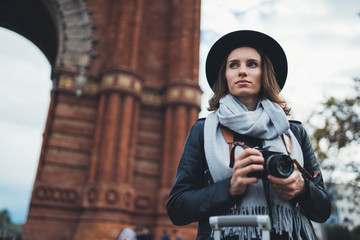 Photographer tourist with retro photo camera. Portait girl in hat travels in Triumphal arch Barcelona. Holiday concept in europe street. Traveler hipster shoot architecture city, copy space mockup