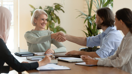 Smiling mature female leader handshake male partner at team meeting