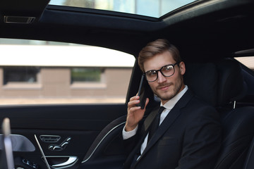 Handsome confident businessman in suit talking on smart phone and working using laptop while sitting in the car.