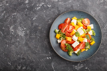 Vegetarian salad with broccoli, tomatoes, feta cheese, and pumpkin on a blue ceramic plate on a black concrete background, top view