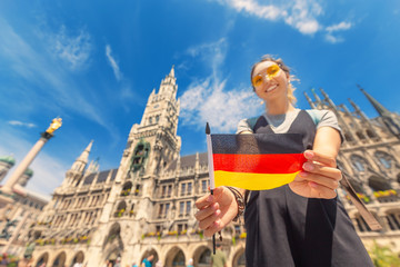 A happy girl with a German flag poses against the background of the city hall in Munich. Travel and immigration to Germany - obrazy, fototapety, plakaty